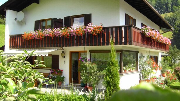 A two-story house with a balcony adorned with flowers, surrounded by lush greenery.