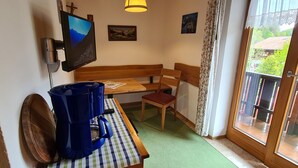 A room with a wooden bench, a table with a checkered tablecloth, a blue vacuum cleaner, a television mounted on the wall, and a window with a view of greenery.