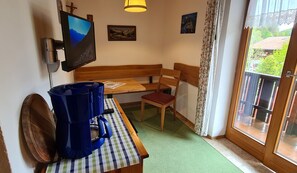 Ein Zimmer mit einer hölzernen Bank, einem Tisch mit kariertem Tischläufer, einem blauen Staubsauger, einem an der Wand montierten Fernseher und einem Fenster mit Blick auf Grünflächen.