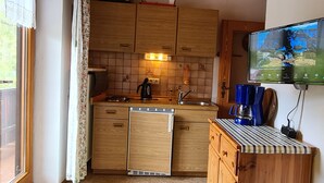 A compact kitchen with wooden cabinets, a built-in dishwasher, a coffee maker, and a television mounted on the wall.