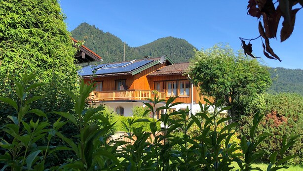 Une maison en bois avec un toit en pente et des panneaux solaires, entourée d’une végétation luxuriante et avec des montagnes en arrière-plan.