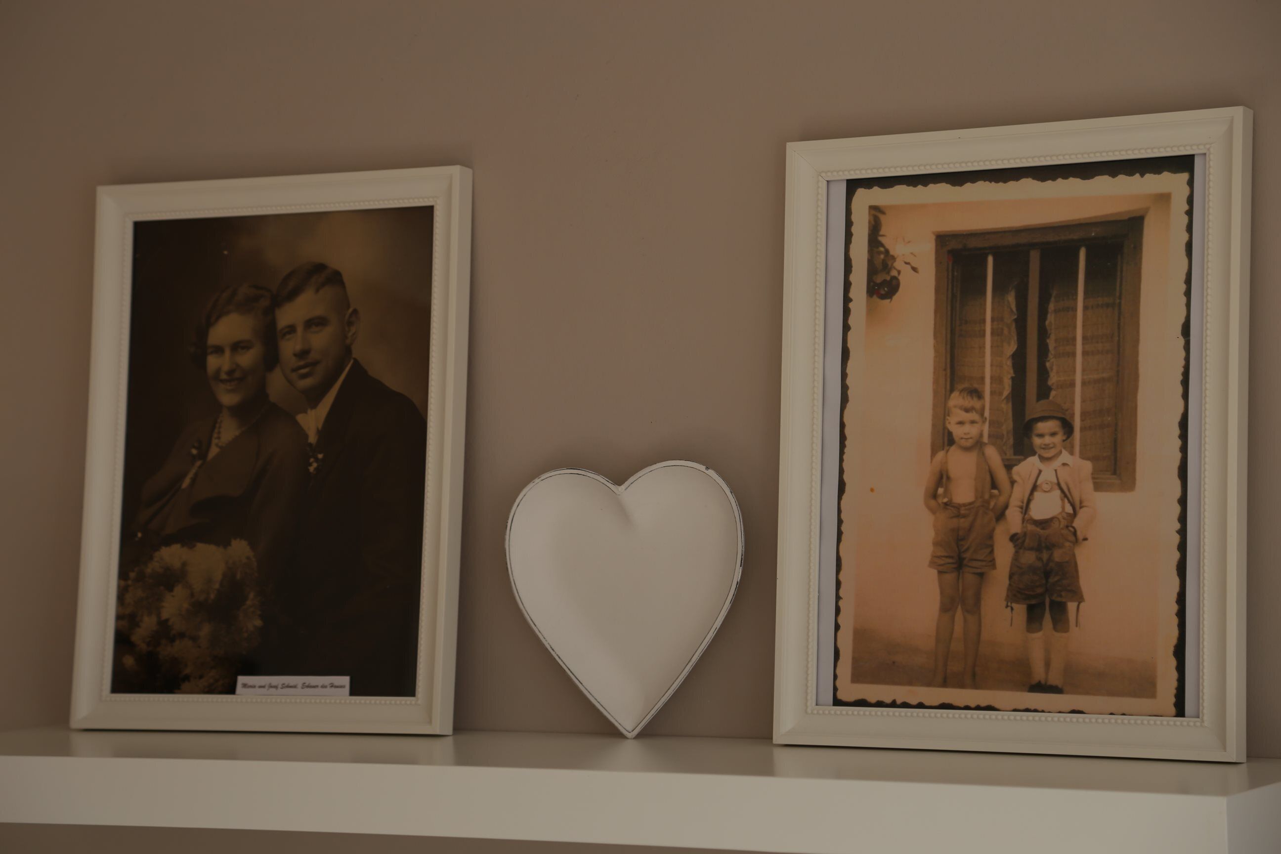 Two framed photographs on a shelf, one of an adult couple and one of two children, with a heart-shaped decoration between them.