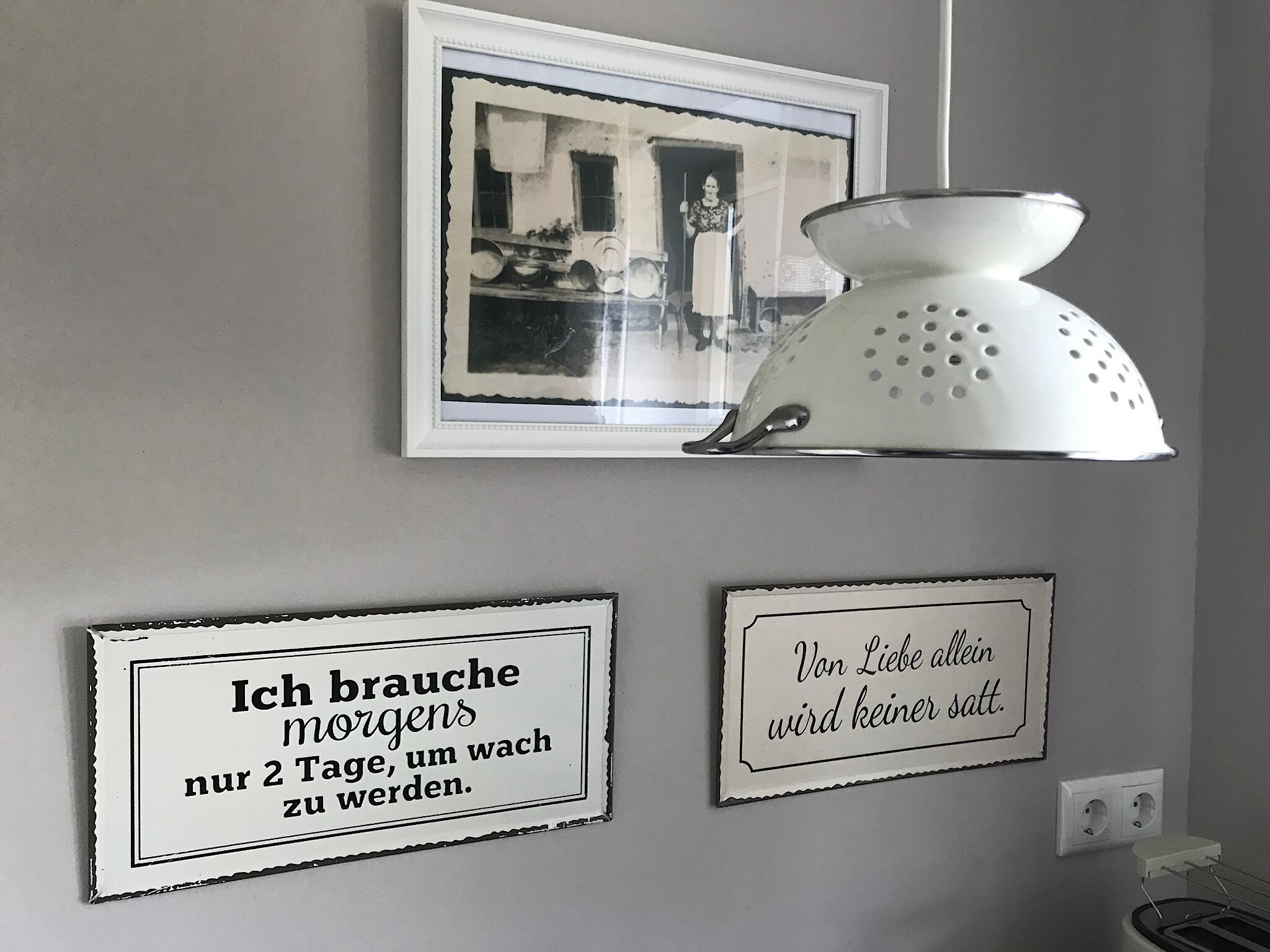 A wall with three framed items: a black and white photo, a white colander, and two signs with German text.