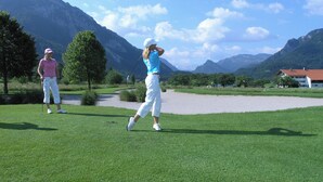 Un campo de golf con montañas al fondo, cielo despejado y un edificio con techo rojo.