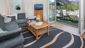 Una sala de estar moderna con sofá, una mesa de centro y televisor. Tiene un balcón con mobiliario exterior y vistas a la montaña.