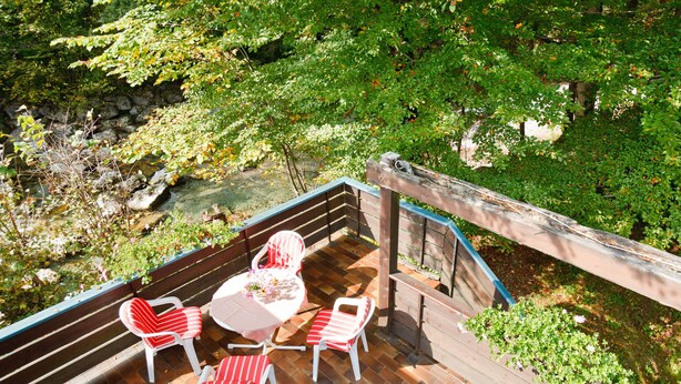 A wooden deck with a table and four striped chairs, surrounded by greenery and a stone wall. - Apartment 2 upper floor 57sqm, balcony, kitchenette, 2 bedrooms, living room (Ruhpolding)