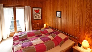 A bedroom with a bed, wooden walls, a nightstand with a lamp, a framed picture, and a window with curtains. - Apartment 2 upper floor 57sqm, balcony, kitchenette, 2 bedrooms, living room (Ruhpolding)