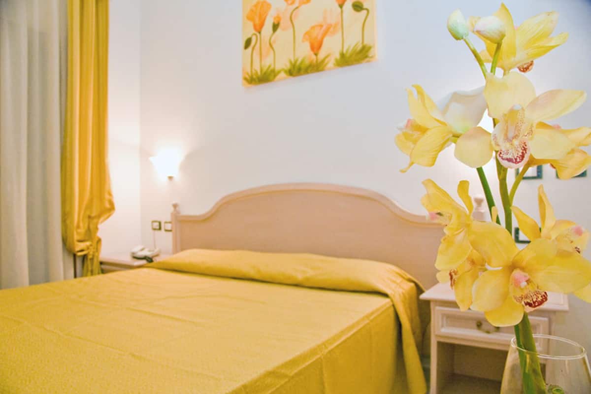 A hotel room with a bed, a nightstand, a vase with flowers, and a painting on the wall.