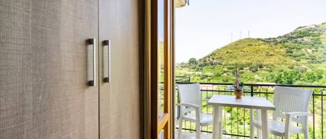 Un balcón con una mesa y sillas, vista a un paisaje verde con colinas, y una puerta de madera con manijas.
