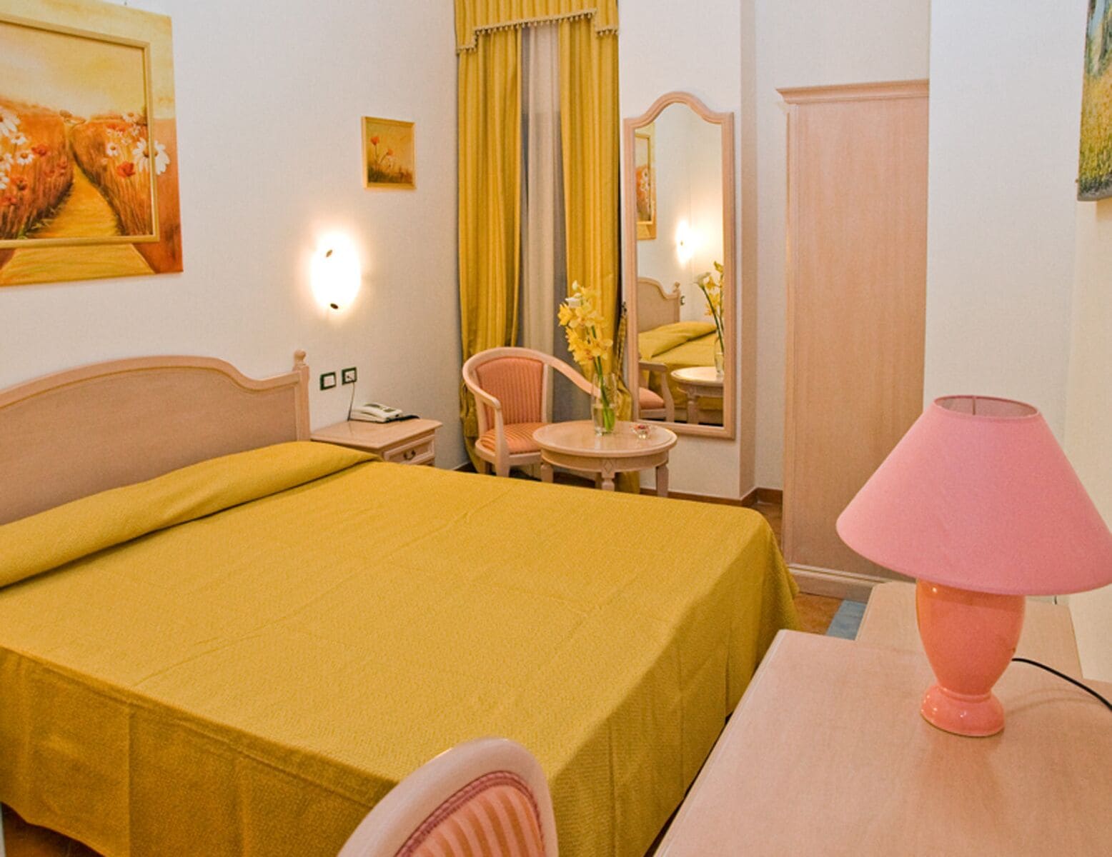 Een hotelkamer met een bed, een stoel, een klein tafeltje, een lamp, een spiegel en een schilderij aan de muur.