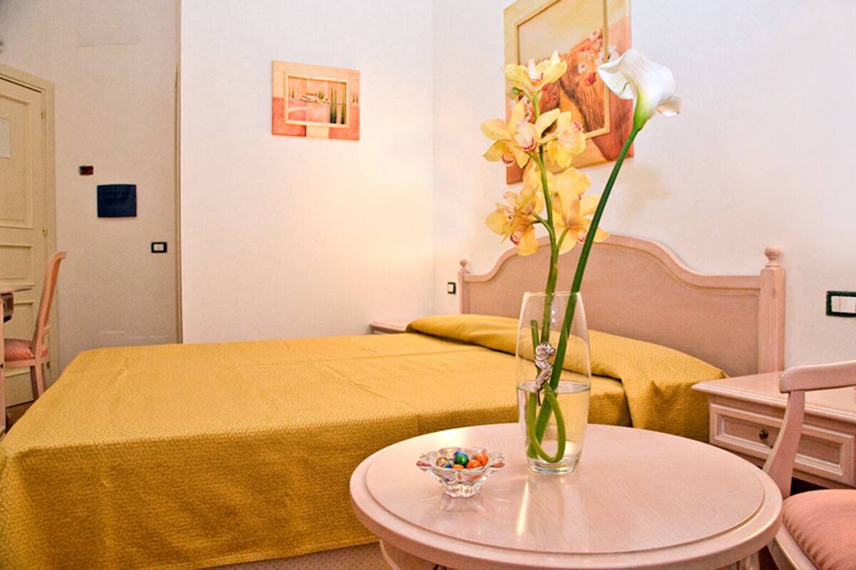 A hotel room with a bed, a bedside table with a vase of flowers, a small table with a bowl of candy, and a chair.
