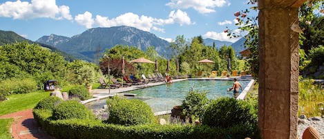 Una piscina con sillas de descanso y sombrillas, rodeada de vegetación y montañas al fondo.
