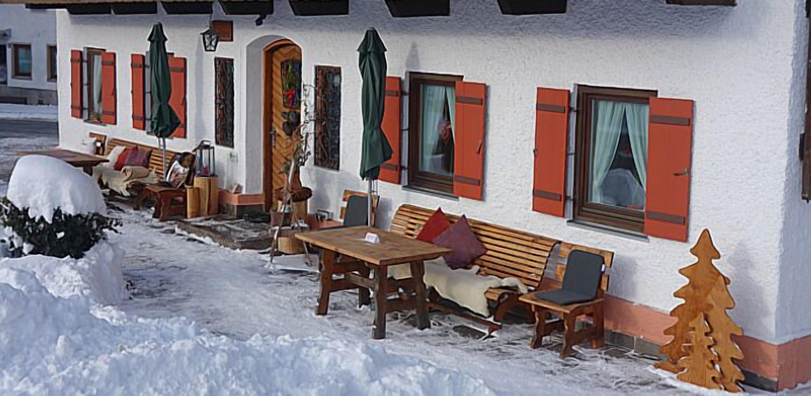 A snow-covered outdoor seating area with wooden tables and benches, a wooden bench with a cushion, and a small Christmas tree.