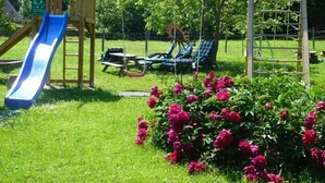 A playground with a slide and swings, surrounded by lush greenery and flowers. - Ferienwohnung Dahoam für 2-6 Personen, 78 qm (Marktschellenberg)
