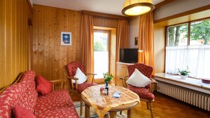A cozy room with wooden walls, a sofa, two armchairs, a table with a tablecloth, a TV, and a window with curtains. - Ferienwohnung -3- (35qm), Terrasse, Küche extra, 1 Schlaf- und 1 Wohnzimmer, max 2 Personen (Ruhpolding)