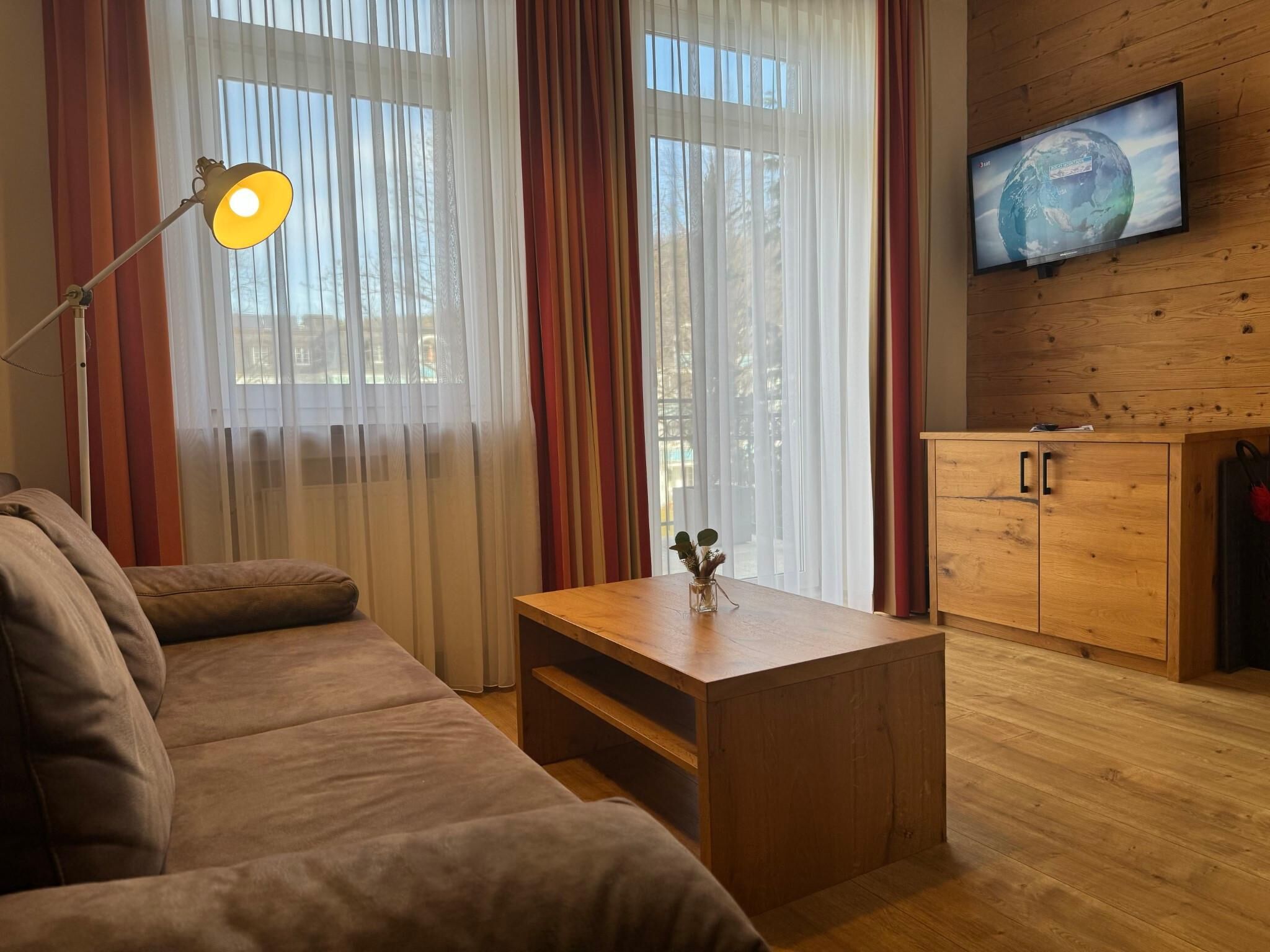 Ein gemütliches Wohnzimmer mit einem Holzsofa, einem kleinen Holztisch mit einer Vase, einer Lampe, einem Fernseher an der Wand und einer Holzkommode.