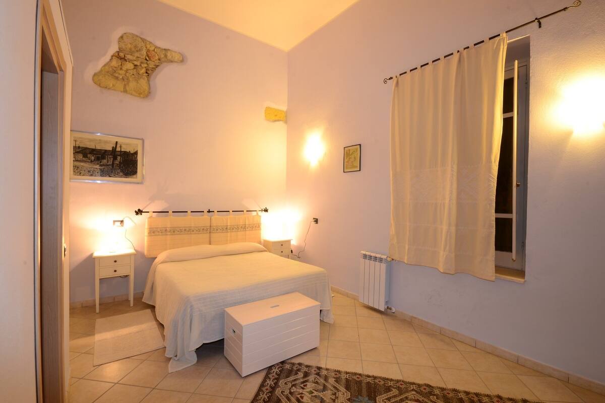 A bedroom with a bed, bedside lamps, a nightstand, a rug, a radiator, and a window with curtains.