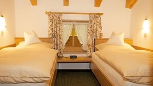 Une chambre double avec deux lits, une tête de lit en bois, une petite table avec une radio et une fenêtre agrémentée de rideaux à motifs floraux.