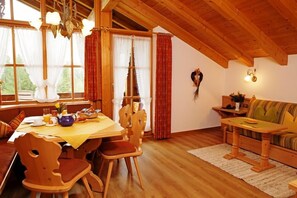 A wooden dining table with chairs, a sofa, and a chandelier in a room with wooden beams and large windows. - (4) Drei-Raum-Ferienwohnung 65qm, 2-6 Personen, Dusche/WC, Wohn-Schlafraum + 2 Extra Schlafzimmer, Küche, großer Balkon (Reit im Winkl)