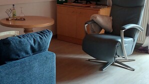 A modern living room with a high ceiling, a black leather chair, a blue sofa, a wooden cabinet, and a round table with a glass. - Apartment- 3 persons, 50sqm, 1 bedroom, balcony, WLAN (Oberwössen)