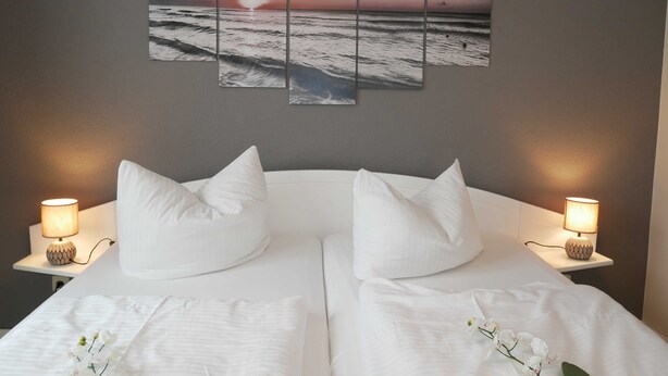 Een tweepersoonsbed met witte lakens en twee kussens, met nachtkastjes en lampen ernaast. Boven het bed hangt een kunstwerk bestaande uit meerdere panelen, waarop een zonsondergang boven de oceaan is afgebeeld.