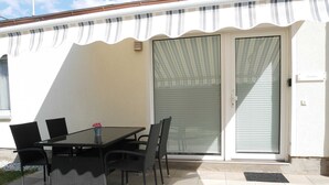 Een terras buiten met een tafel, stoelen, een schuifdeur en een witte zonnescherm.