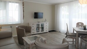 Een woonkamer met een televisie, een wit kastje, een beige bank, een glazen salontafel, en een eethoek met een tafel en stoelen.