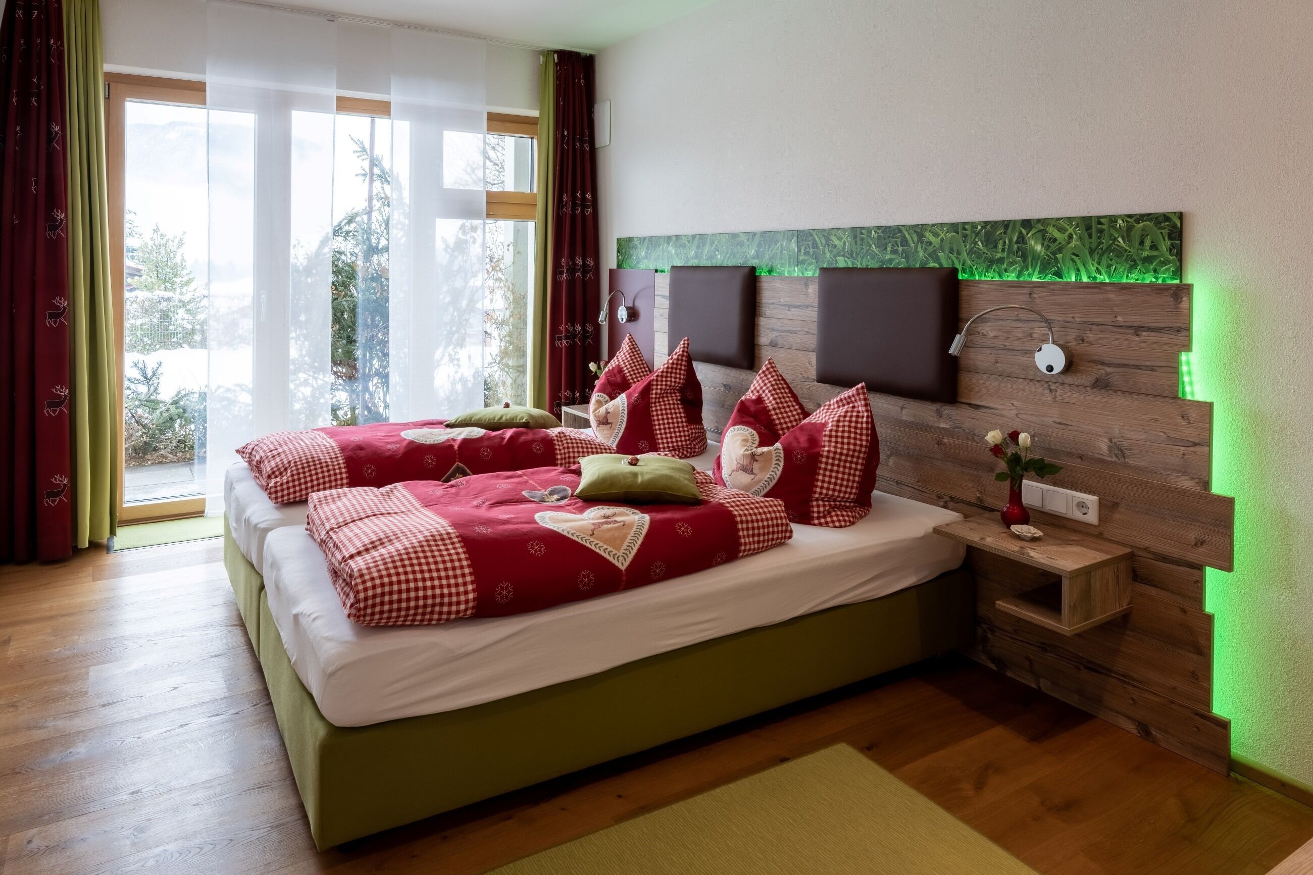 A double bed with red and white checkered bedding, a wooden headboard with green LED lighting, a bedside table with a vase, and a view of greenery outside.