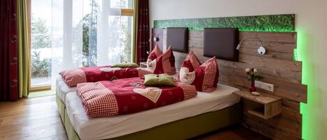 A double bed with red and white checkered bedding, a wooden headboard with green LED lighting, a bedside table with a vase, and a view of greenery outside.