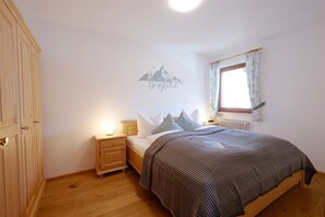 A bedroom with a bed, nightstand, wooden wardrobe, wall art, window with curtains, and a radiator.