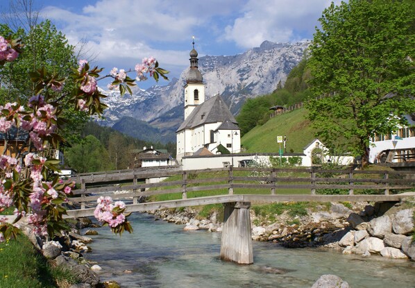 Un puente de madera sobre un río transparente, una iglesia con campanario y montañas nevadas al fondo.