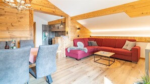 A cozy living room with a red sofa, grey chairs, a wooden ceiling, and a small dining table. - Three-room apartment (8) Haus Resi, 70sqm, two bedrooms, two bathrooms, living room, kitchen, balcony (Reit im Winkl)
