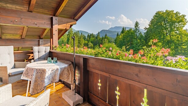 A wooden deck with outdoor furniture, a table with a patterned tablecloth, and a view of a mountainous landscape with trees and flowers. - Three-room apartment (8) Haus Resi, 70sqm, two bedrooms, two bathrooms, living room, kitchen, balcony (Reit im Winkl)