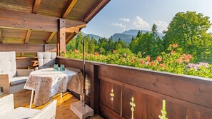 Een houten terras met tuinmeubilair, een tafel met een tafelkleed met dessin en een uitzicht op een bergachtig landschap met bomen en bloemen.