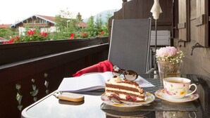 Una mesa con una porción de pastel, una taza de café y un libro en un balcón con barandillas de madera y vistas a edificios y árboles.
