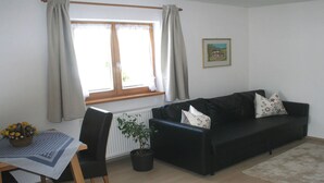 A living room with a black sofa, a table with a basket of flowers, a radiator, and a window with curtains. - (2) Ferienwohnung 36m², 2 Personen mit seperaten Schlafzimmer, Du/WC, Balkon (Reit im Winkl)