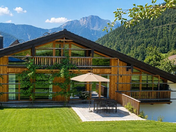 A two-story wooden house with large windows, a balcony, and a patio with outdoor furniture.