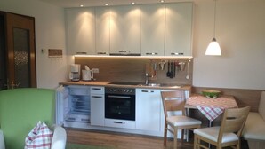 A compact kitchen with white cabinets, a built-in oven, and a coffee maker. There is a dining area with a table and chairs, and a green carpet. - Ferienwohnung Nr. 2 Kehlsteinblick, 1 bis 2 Personen, 45 qm, 1 Schlafzimmer, Wohnküche und Balkon (Schönau am Königssee)