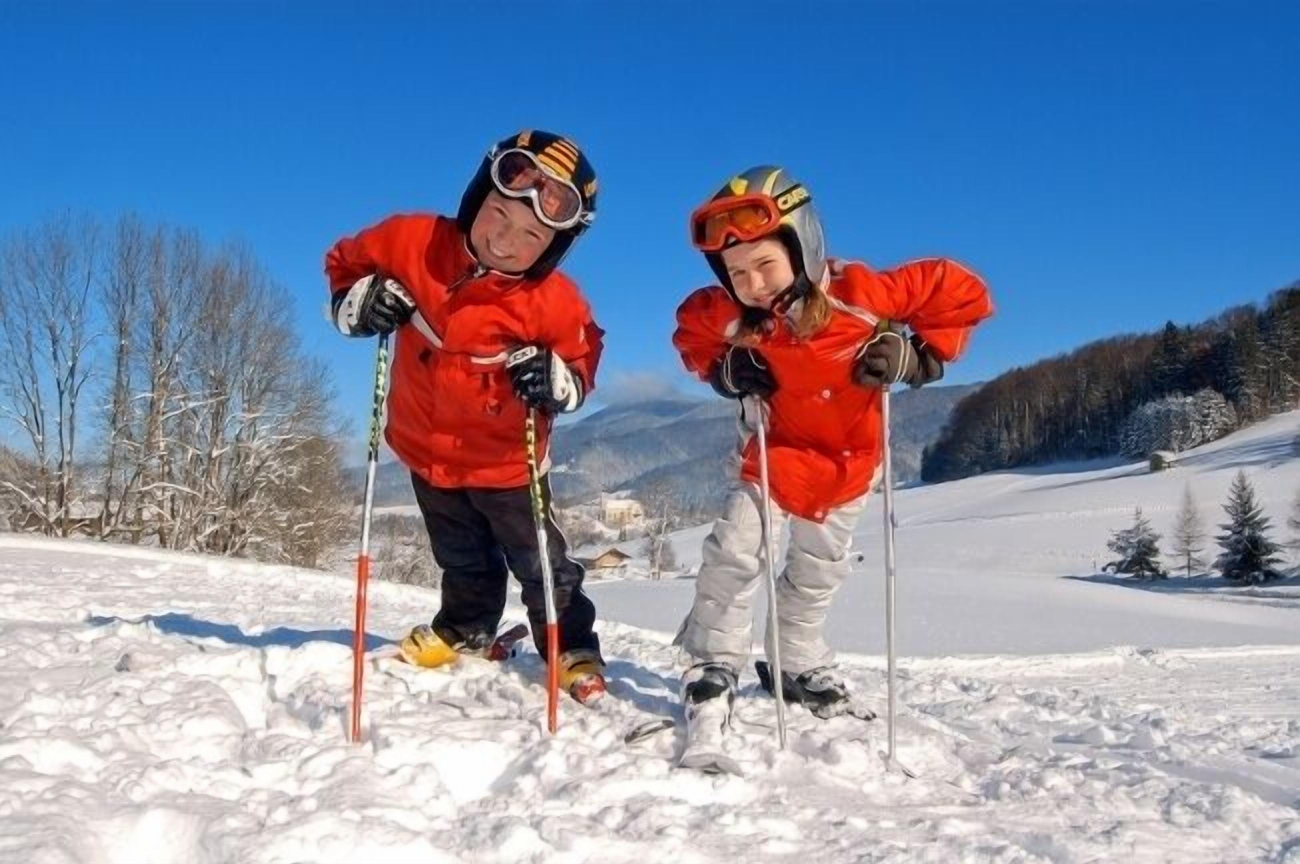 Two people skiing on a snowy slope with trees and mountains in the background.