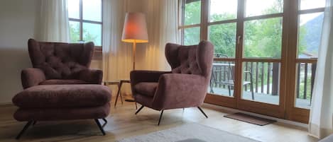 A room with two armchairs, a table, and a lamp. There are large windows with a view of greenery outside.