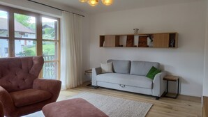 Un salón con un sofá gris, un sillón marrón, una estantería de madera con objetos decorativos y vistas a una casa a través de ventanas grandes.