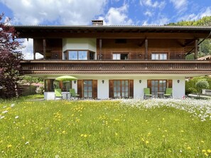 Una casa a due piani in legno con un balcone, un portico coperto con posti a sedere all'aperto e un giardino con fiori gialli.