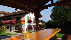Et træbord med et glas og en lille orange beholder, foran en hvid bygning med altaner pyntet med farverige blomster.