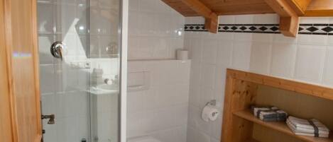 A bathroom with a glass shower enclosure, a toilet, a sink, and a wooden shelf with towels.