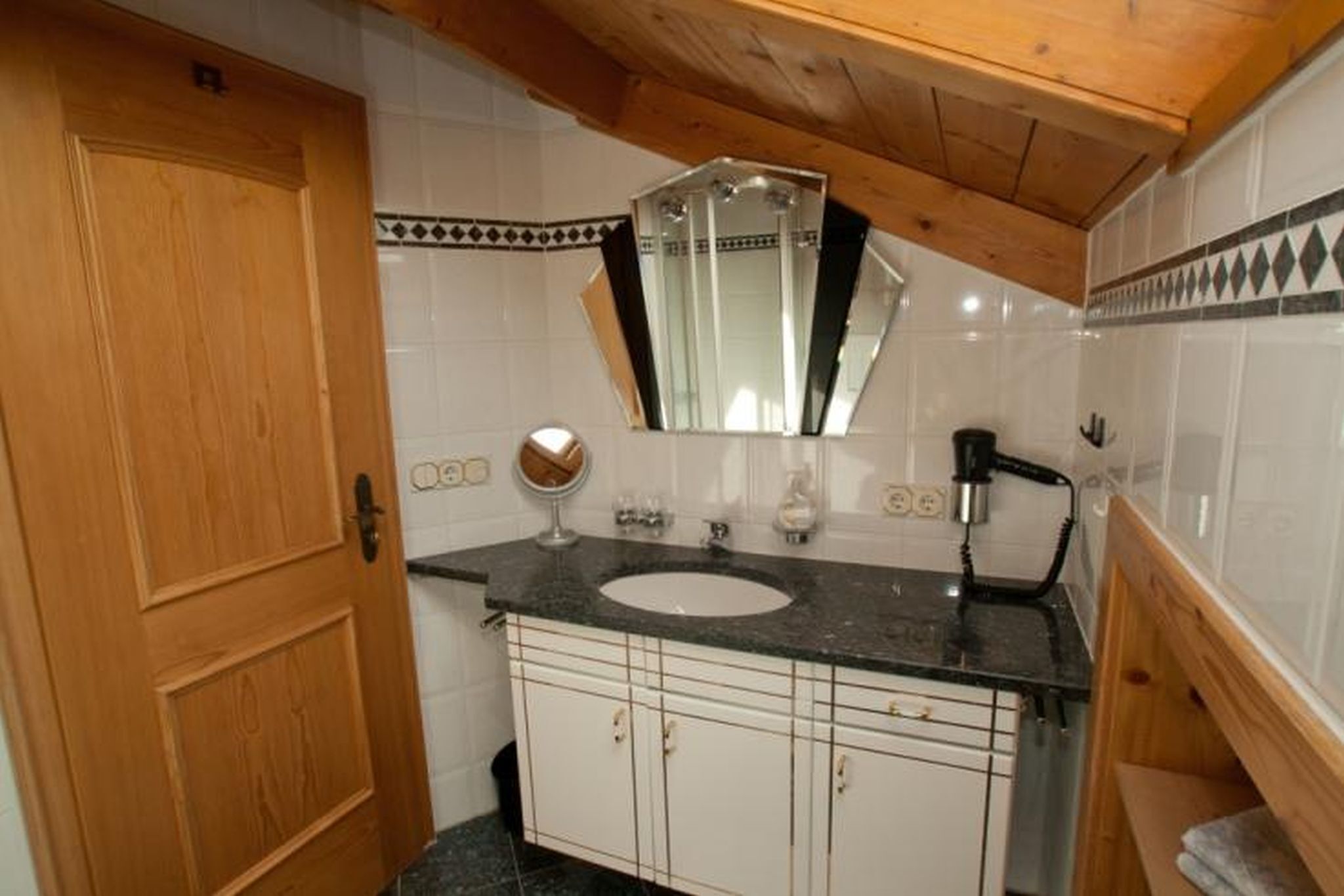 Une salle de bain avec une porte en bois, des murs recouverts de carreaux blancs, un lavabo sur un plan de travail noir, un miroir et un porte-serviettes.