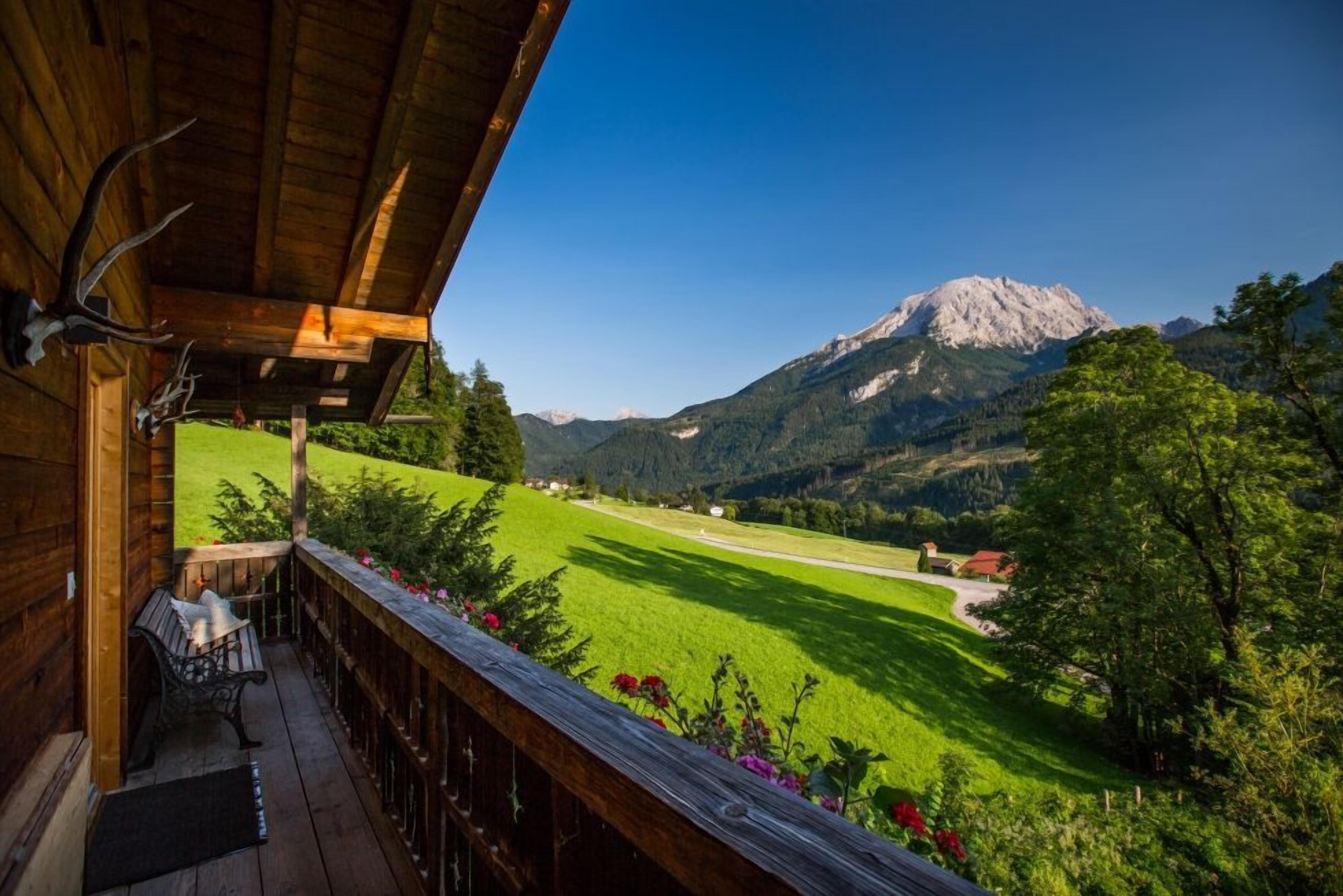 Un balcón de madera con un banco, con vista a un campo verde y una montaña.