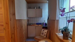 Ein kleiner, gemütlicher Raum mit Holzwänden und einer Küchenzeile. Ein kleiner Tisch mit einer Fernbedienung und einem Kissen ist vorhanden. Ein Fenster mit violetten Orchideen ist sichtbar.