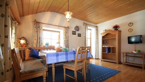 A cozy dining area with a wooden table, chairs, and a blue rug. There is a television on a wooden stand and a bookshelf with various items. - Ferienwohnung 1 "Hortensie" (Nußdorf i. Chiemgau)