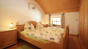 A bedroom with a wooden ceiling, a bed with floral bedding, a nightstand with a lamp, a window with curtains, and a radiator. - Ferienwohnung 1 "Hortensie" (Nußdorf i. Chiemgau)