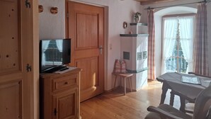 Ein gemütliches Zimmer mit Holzmöbeln, einem Fernseher auf einem Schrank, einem kleinen Tisch und einem Fenster mit Vorhängen.